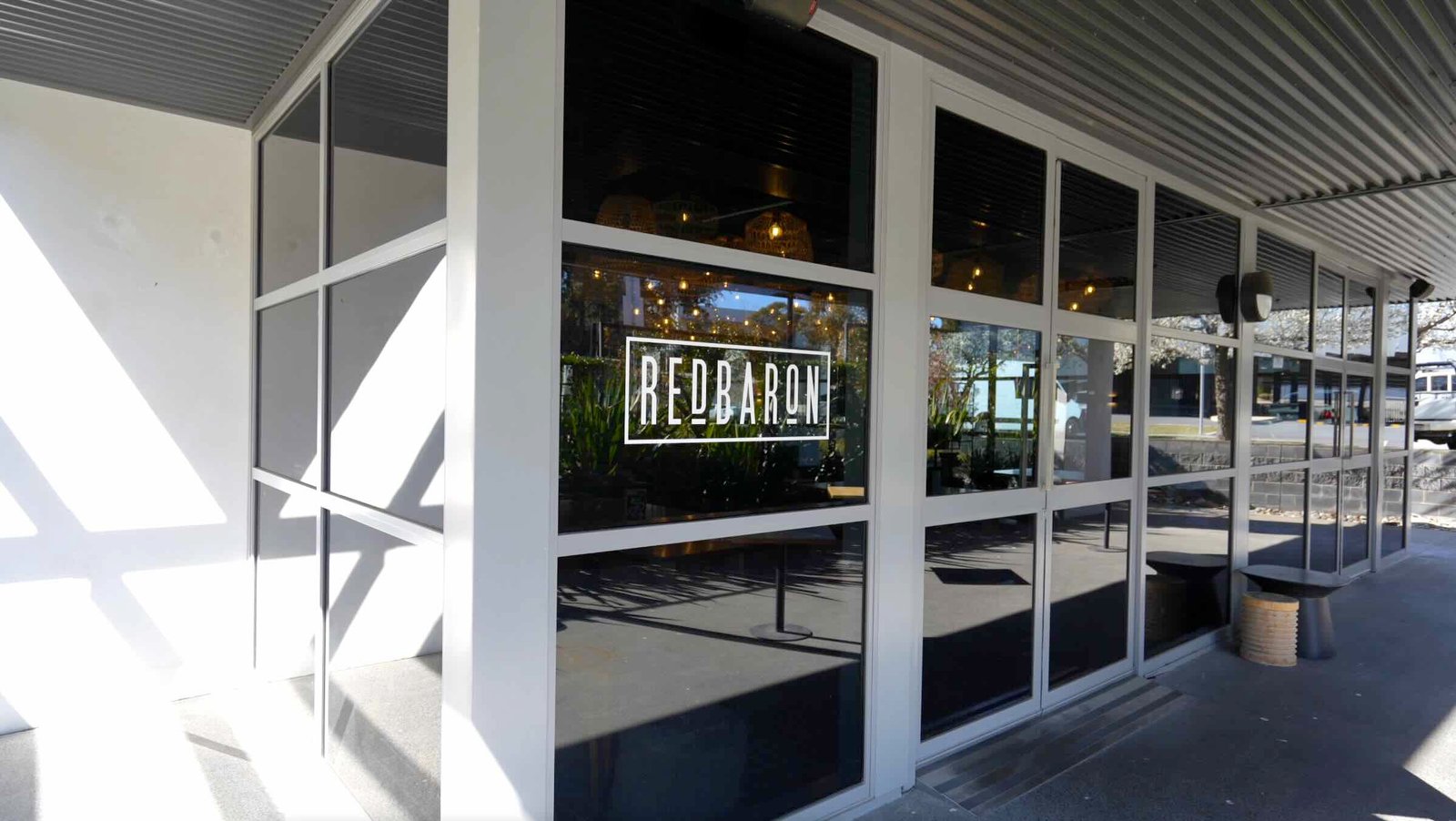 Red Baron cafÃ© - covered outdoor pergola terrace with timber beams, festoon lights and views to the precinct lawn and bronze sculpture
